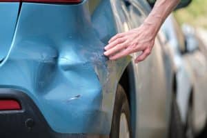 Driver hand examining dented car with damaged fender parked on city street side. Road safety and vehicle insurance concept.