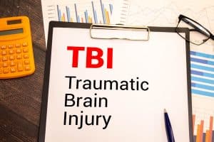 Clipboard with the text "TBI Traumatic Brain Injury" surrounded by charts, a pen, calculator, and eyeglasses, representing medical or legal analysis.