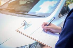 Person filling out documents on a clipboard on a car hood, outdoors in daylight.